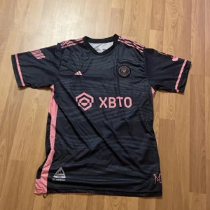 Svart och rosa fotbollströja med Messi. Brasilianska landslaget tröja  -  Bjud me ett pris.  Snygg svart fotbollströja med rosa detaljer och Messi-tryck på ryggen. Tröjan har korta ärmar och är perfekt för fotbollsfans. Adidas-logga och sponsortryck på framsidan. Brasilianska tröja 