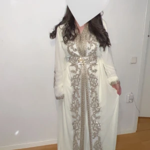 Vit kaftan med gulddetaljer - Elegant vit kaftan med vackra gulddetaljer och broderier. Klänningen har långa ärmar och en v-ringad design med ett bälte i midjan som framhäver figuren. Perfekt för speciella tillfällen. Storlek passar både S och M. Använd endast i några timmar.