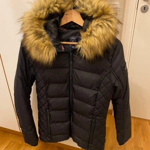 Svart dunjacka med pälskrage från Polo Club - Snygg svart dunjacka från Polo Club med en fluffig pälskrage i beige. Jackan har en quiltad design och dragkedja framtill. Perfekt för kyliga dagar.