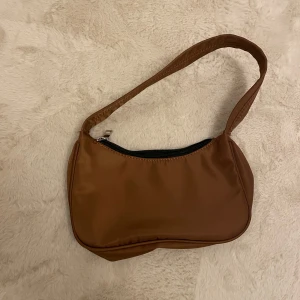 Brun axelväska med dragkedja - Snygg brun axelväska i syntetmaterial med en praktisk dragkedja upptill. Perfekt för att bära dina nödvändigheter med stil. Enkel och stilren design som passar till många tillfällen. Ingen prislapp på, men aldrig använts.