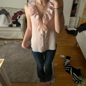 Rosa ärmlös blus med volang - Säljer en söt rosa blus med volangdetaljer framtill. Blusen är ärmlös och har en v-ringad design som ger en feminin touch. Perfekt för varmare dagar eller som ett lager under en kavaj.