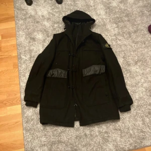 Svart rock från Stone Island - Säljer en svart rock från Stone Island med huva och quiltat foder. Jackan har långa ärmar och en klassisk design med knappar och dragkedja. Perfekt för kyligare dagar. Storlek S-M