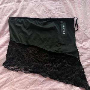Svart topp med spetsdetalj från Motel - Snygg svart kjol från Motel med asymmetrisk design och spetsdetalj. Perfekt för att ge en edgy touch till din outfit. Toppen har en tight passform och är gjord i ett mjukt material som sitter skönt på kroppen.