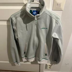 Grå jacka från Adidas - Snygg  jacka från Adidas med dragkedja och gröna detaljer. Jackan har långa ärmar och två fickor med dragkedjor. Perfekt för en sportig look.