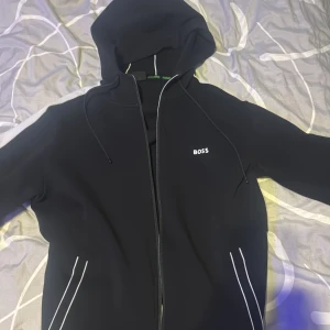 Hugo boss dress  - Snygg svart hoodie från Boss med dragkedja och huva. Den har vita detaljer och snören vid huvan. Pch matchande byxor väldigt bra skick, ny pris cirka 3000 för byxor och tröja storlek L men passar m