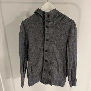 Grå cardigan med luva - Säljer en stilren grå cardigan/hoodie med knappar framtill. Tröjan har en huva och långa ärmar, perfekt för kyligare dagar. Den är i en neutral grå färg som passar till det mesta.