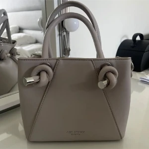 Beige handväska - Elegant svart  handväska  med unika knutdetaljer på handtagen. Väskan har en stilren design och är perfekt för att bära dina nödvändigheter med stil. Den är kompakt men rymlig nog för det viktigaste. Väskan är i svart!! Inte beige 