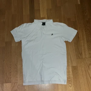 Vit kortärmad pikétröja - Säljer en stilren vit pikétröja med korta ärmar och en liten broderad logotyp på bröstet. Perfekt för en avslappnad och snygg look.