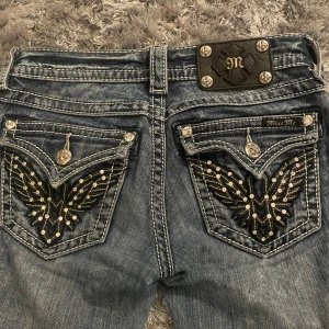 Blå jeans från Miss Me - Snygga blå jeans från Miss Me med unika broderade detaljer och nitar på bakfickorna. De har en lågmidjad passform och är bootcut. Jeansen har slitna detaljer men vid sömnen där de går ner över hälen är det däremot slitna. Utöver det är det i extremt bra skick.                                     Dessa är i strl w24 men jag som är 170 lång kommer i de, däremot väldigt små midjan. 