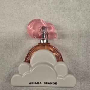 Ariana Grande Cloud Pink Parfym - Charmig parfymflaska med ett rosa molnformat lock och en bas i form av ett vitt moln. Doften är en del av Ariana Grandes populära kollektion. Förpackningen är i en glansig rosa kartong med texten 'Cloud Pink'. Perfekt för den som älskar söta och fruktiga dofter.