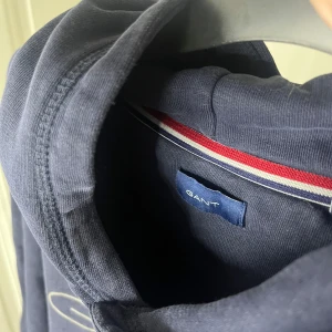 Mörkblå hoodie från GANT - Säljer en snygg mörkblå hoodie från GANT med logga och emblem på bröstet. Tröjan har en klassisk känguruficka och en bekväm huva. Perfekt för en avslappnad stil.