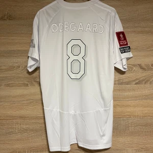 FA Cup Arsenal Ødegaard XL - Snygg vit Arsenal fotbollströja från Adidas med Ødegaard och nummer 8 på ryggen. Tröjan har korta ärmar och flera tryck, inklusive Emirates FA Cup och 'No room for racism'. Perfekt för fans av Arsenal och Ødegaard! Storlek XL