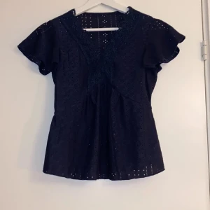 Mörk blå blus med spetsdetaljer - Elegant svart blus med spetsdetaljer och korta ärmar. Perfekt för en stilren look med v-ringad design och volangdetaljer vid ärmarna. Köpt här på plkck.