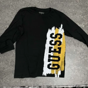 Svart långärmad tröja från Guess - Snygg svart långärmad tröja från Guess med ett stort tryck i vitt och gult på framsidan. Perfekt för en avslappnad stil med en touch av färg.
