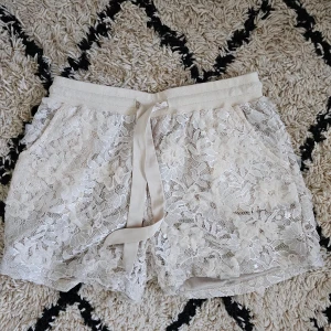 Beige spets shorts från Buch  Copenhagen  - Snygga beige shorts i spets. De har en elastisk midja med knytband och ett vackert blommigt mönster. Perfekta för en somrig look!