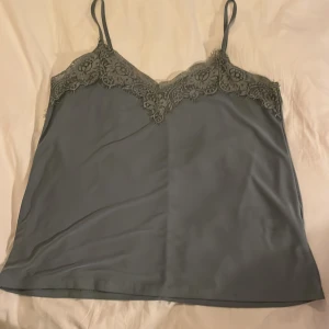 Grönt linne med spetsdetaljer - Säljer ett stilrent grönt linne med tunna axelband och vacker spetsdetalj längs halsringningen. Perfekt för en elegant och avslappnad look.