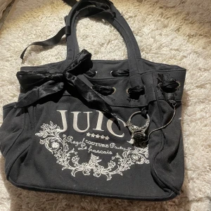 Svart väska från Juicy Couture - Snygg svart väska från Juicy Couture med dekorativa detaljer och tryck. Det på kanten åker av lite och det är någon droppe nagellim på baksidan men det är inget man märker av. Väskan har en rosett och ett hjärta med 'J' på baksidan. Insidan är fodrad med ett rosa mönster av kronor och text. Perfekt för att ge en touch av stil till din outfit. ((Pris kan diskuteras))