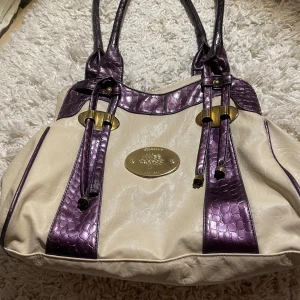 Beige och lila axelväska med leopardmönster - Snygg axelväska i beige med lila krokodilmönstrade detaljer och leopardmönstrat foder. Väskan har dubbla handtag och en dekorativ metallplatta framtill. Perfekt för att ge din outfit en unik touch!