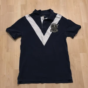 Mörkblå pikétröja från Ralph Lauren - Säljer en snygg mörkblå pikétröja från Ralph Lauren med vit V-formad detalj och broderad logotyp på bröstet. Perfekt för en stilren look. Den är i storlek L/G (14-16)!