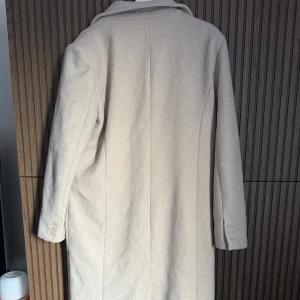 Beige kappa från NA-KD - Snygg beige kappa från NA-KD med klassisk design och knappar framtill. Lång med slits. Litte nopprig men går att få bort utan problem 
