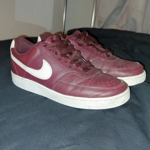 Vinröda Nike sneakers - Säljer dessa snygga vinröda Nike sneakers med vit sula och vit swoosh. Skorna har snörning och är tillverkade i skinn. Perfekta för en casual look.  Storleken är 39. Nypris ligger runt 700-800. Mitt pris 399. Dem är lite creasade och har några små trådar som sticker ut, annars bra skick. Priset är ej hugget i sten. Om du har några frågor eller tankar så skriv gärna😊
