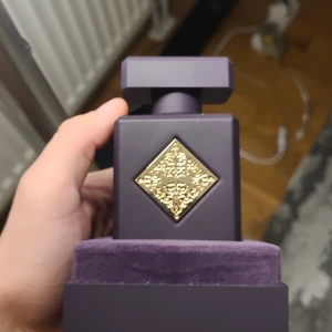 Lila parfymflaska med gulddetalj - Elegant parfymflaska i en djup lila nyans med en fyrkantig form. Flaskan har en dekorativ gulddetalj i form av en diamant på framsidan. Den kommer i en matchande lila förpackning med sammetsliknande yta.