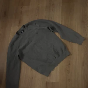 Grå stickad tröja från Gant - Säljer en stilren grå stickad tröja från Gant med rund halsringning och ett diskret broderat emblem på bröstet. Perfekt för kyligare dagar.