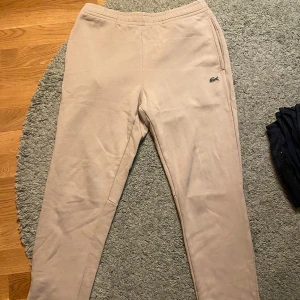Beige mjukisbyxor från Lacoste -   Säljer ett par snygga och stilrena Lacoste-joggers i beige färg. 	•	Storlek: FR 4 (US Medium). 	•	Kvalitet: Mjuk och bekväm bomull, perfekt för både vardagsbruk och avslappning. 	•	Detaljer: Klassisk Lacoste-logga broderad på sidan för en exklusiv look. 	•	