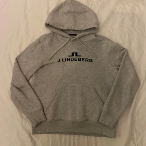 Grå hoodie från J.Lindeberg - Snygg grå hoodie från J.Lindeberg med svart logotyp på bröstet. Hoodien är i bra skick, men några bokstäver är lösa i kanterna. Annars är det inga andra defekter som jag vet om. Skriv i dm om frågor!