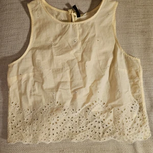 Gul ärmlös topp från H&M Divided - Säljer en söt gul ärmlös topp från H&M. Divided i storlek 38 med broderade detaljer och knytning i ryggen. Toppen har ett vackert hålmönster längs nederkanten som ger en romantisk touch.
