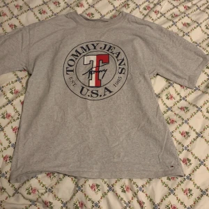  Oversized grå t-shirt från Tommy Jeans (Hilfiger)  - Säljer en grå oversized t-shirt från Tommy Jeans med ett stort tryck på framsidan. T-shirten har korta ärmar och en rund halsringning. Perfekt för en avslappnad stil.