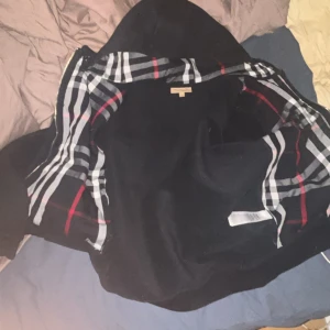 Svart hoodie från Burberry - Snygg svart hoodie från Burberry med klassiskt rutigt mönster på insidan av huvan. Jackan har dragkedja framtill och är tillverkad i mjukt material för extra komfort. Perfekt för en stilren look.