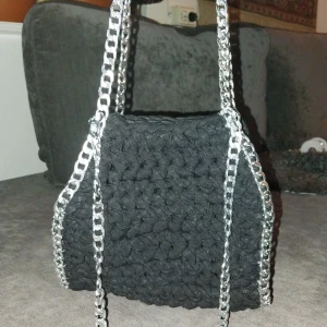 Svart virkad handväska med kedjedetaljer - Snygg svart virkad handväska med silverfärgade kedjor. Perfekt för att ge en edgy touch till din outfit. Väskan har en unik design med en robust känsla.