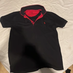 svart och röd pike tröja från polo ralph lauren - Snygg svart pikétröja från Ralph Lauren med röd krage och detaljer. Den har korta ärmar och en liten röd logotyp på bröstet. Perfekt för en stilren look.
