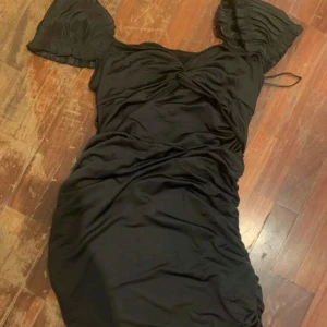 Svart klänning med rynkade detaljer - Elegant svart klänning med rynkade detaljer och korta ärmar. Den har en v-ringad design och en tight passform som framhäver figuren. Perfekt för en kväll ute.