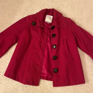 Röd jacka från Bershka - Snygg röd jacka från Bershka med stora svarta knappar. Jackan har en klassisk krage som går att knäppa uppe eller behålla nere. Jackan har trekvartsärmar. Perfekt för höst och vår.