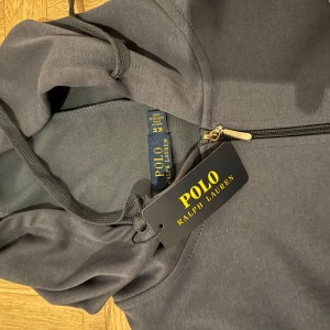 Blå huvtröja från Polo Ralph Lauren - Säljer en helt ny snygg blå huvtröja från Polo Ralph Lauren med dragkedja och det klassiska logotypbroderiet på bröstet. Tröjan har långa ärmar och en praktisk huva med dragsko. Perfekt för en avslappnad stil.