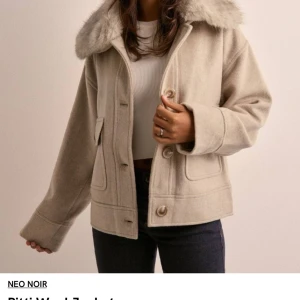 Beige jacka med pälsdetaljer från Neo Noir - Snygg beige jacka från Neo Noir med mjuk pälsdetalj på kragen. Jackan har knappar framtill och praktiska fickor. Perfekt för kyligare dagar.