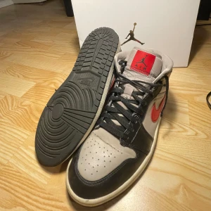 Air Jordan 1s Svart, Grå, Röd - Snygga Nike Air Jordans i svart och grått med röda detaljer. Skorna har en klassisk design med snörning och perforerad tå för extra ventilation. Perfekta för både stil och komfort.