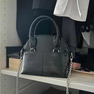 Svart krokodilpräglad handväska - Snygg svart handväska med krokodilpräglat mönster och silverdetaljer. Väskan har en kedja som axelrem och två handtag för olika bäralternativ. Perfekt för att ge en elegant touch till din outfit.