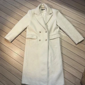 Vit kappa med knappar - Elegant vit kappa med dubbelknäppning och klassisk krage. Perfekt för kyligare dagar med sin stilrena design och långa ärmar. Ett tidlöst plagg som passar till många tillfällen.