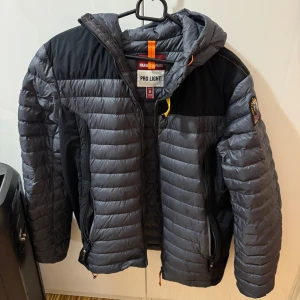 Grå dunjacka från Parajumpers - Säljer en grå dunjacka från Parajumpers med huva och dragkedja. Jackan har en broderad logga på ärmen och orangea detaljer. Perfekt för kyliga dagar.