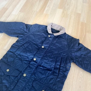 Blå quiltad jacka från Barbour - Snygg blå quiltad jacka från Barbour med knappar och en kontrasterande krage i beige manchester. Den här jackan har varit en riktig räddning för mig kalla vår och höst dagar. 