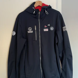 Marinblå jacka från Marine Scandinavian Sportswear - Snygg marinblå jacka från Marine Scandinavian Sportswear med broderade detaljer och dragkedja. Jackan har en huva och är tillverkad i 100% polyester. Perfekt för svala dagar.