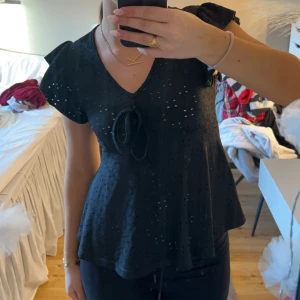 Svart blus med volangärmar - Snygg svart blus med korta volangärmar och knytning framtill. Perfekt för en stilren look med en touch av elegans. Blusen har en lätt glansig yta och en v-ringad halslinje. Står ingen storlek men skulle säga s/m