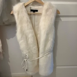 Vit pälsväst från Amisu - Säljer en elegant vit pälsväst från Amisu. Västen har en stilren design med ett knytband i midjan för en justerbar passform. Bandet går att ta av helt❤️krämvit färg men mer mot de vita hållet än de beiga