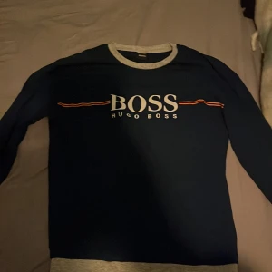 Mörkblå tröja från Hugo Boss - Säljer en snygg mörkblå tröja från Hugo Boss med gråa detaljer vid hals och nederkant. Tröjan har långa ärmar och ett stilrent tryck med märkesnamnet på bröstet. Perfekt för en avslappnad stil.