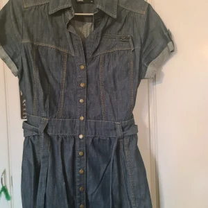 Jeansklänning från Cubus - Snygg jeansklänning från Cubus med korta ärmar och knappar framtill. Klänningen har en klassisk krage och ett bälte i midjan för en fin passform. Perfekt för en avslappnad stil.