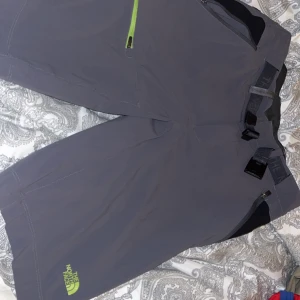 Grå shorts från The North Face - Säljer ett par grå shorts från The North Face i storlek M. De har en cool grön dragkedja på fickan och är perfekta för utomhusaktiviteter. Materialet känns slitstarkt och bekvämt, perfekt för sommarens äventyr. Passformen är loose, vilket gör dem sköna att röra sig i.