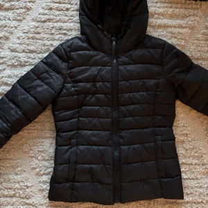Svart pufferjacka från ONLY - Säljer en svart pufferjacka från ONLY. Den är perfekt för kyliga dagar med sin quiltade design och dragkedja framtill. Jackan har en huva och långa ärmar
